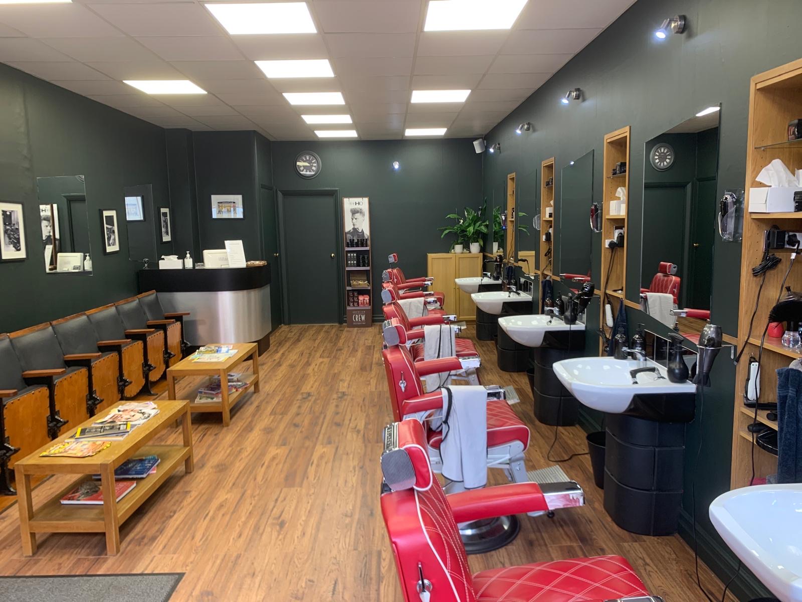 Inside Sweeny&rsquo;s: five red leather barber chairs down one side, the waiting bench and magazines on the other, dark green walls and a wooden floor.
